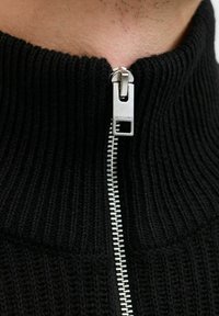 Sort strikket sweater med høj krave og fuld lynlås. Lynlåsen har en sølvfinish med logo-detalje. Struktureret ribbet mønster.