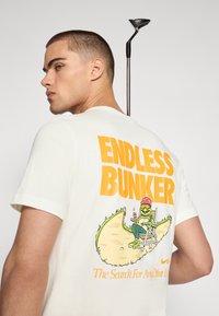 Camiseta de algodón blanca con un colorido estampado gráfico en la parte posterior que incluye el texto "BUNKER INFINITO" y un personaje de dibujos animados bebiendo un trago.