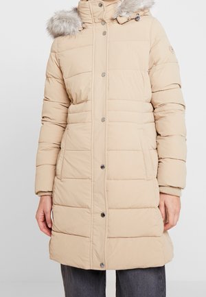 Vrouw draagt een beige doorgestikte lange winterjas met een capuchon met bontkraag en drukknopen, gecombineerd met een donkere broek.