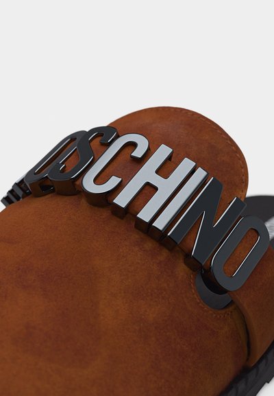 Sandale mule en daim marron avec de grandes lettres en relief noires et argentées formant le mot "MOSCHINO" sur la bride.