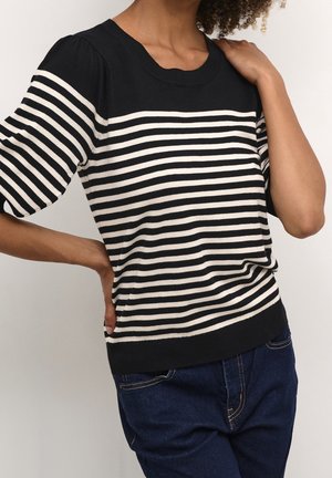 Persoon die een zwart-wit gestreepte korte mouw trui draagt met donkerblauwe jeans, één hand op de heup en de andere die de schouder aanraakt.