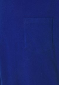 En solid blå tygbit med en sydd rektangulär bröstficka centrerad i rutan.