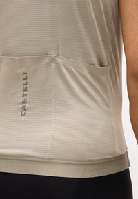 Μπεζ αθλητικό τοπ με υφή, με πίσω τσέπες και το λογότυπο "CASTELLI" στο πίσω μέρος, φορεμένο με μαύρο κάτω μέρος.