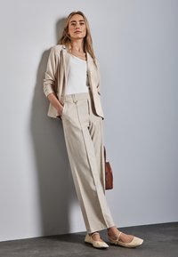 Beige bleiseri valkoisen paidan päällä, vaaleanbeige leveälahkeiset housut, kermaiset kengät ja ruskea laukku. Sileä pinta ja yksinkertaiset muotoiluyksityiskohdat.