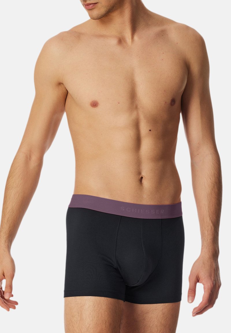 Boxer neri con elastico viola, caratterizzati da un logo. Il tessuto appare liscio con una vestibilità aderente, progettato per comfort e supporto.
