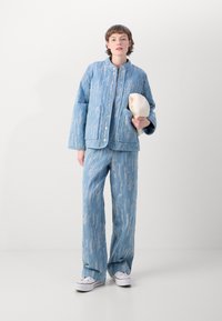 Giacca trapuntata azzurro chiaro con colletto rialzato, abbinata a pantaloni coordinati con un motivo testurizzato, indossati con sneakers bianche.