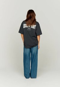 Γυναίκα με μακριά μαλλιά στέκεται μπροστά σε ένα γκρίζο τοίχο, φορώντας μια μαύρη φαρδιά μπλούζα με λευκό κείμενο και μπλε τζιν με φαρδιά πόδια.