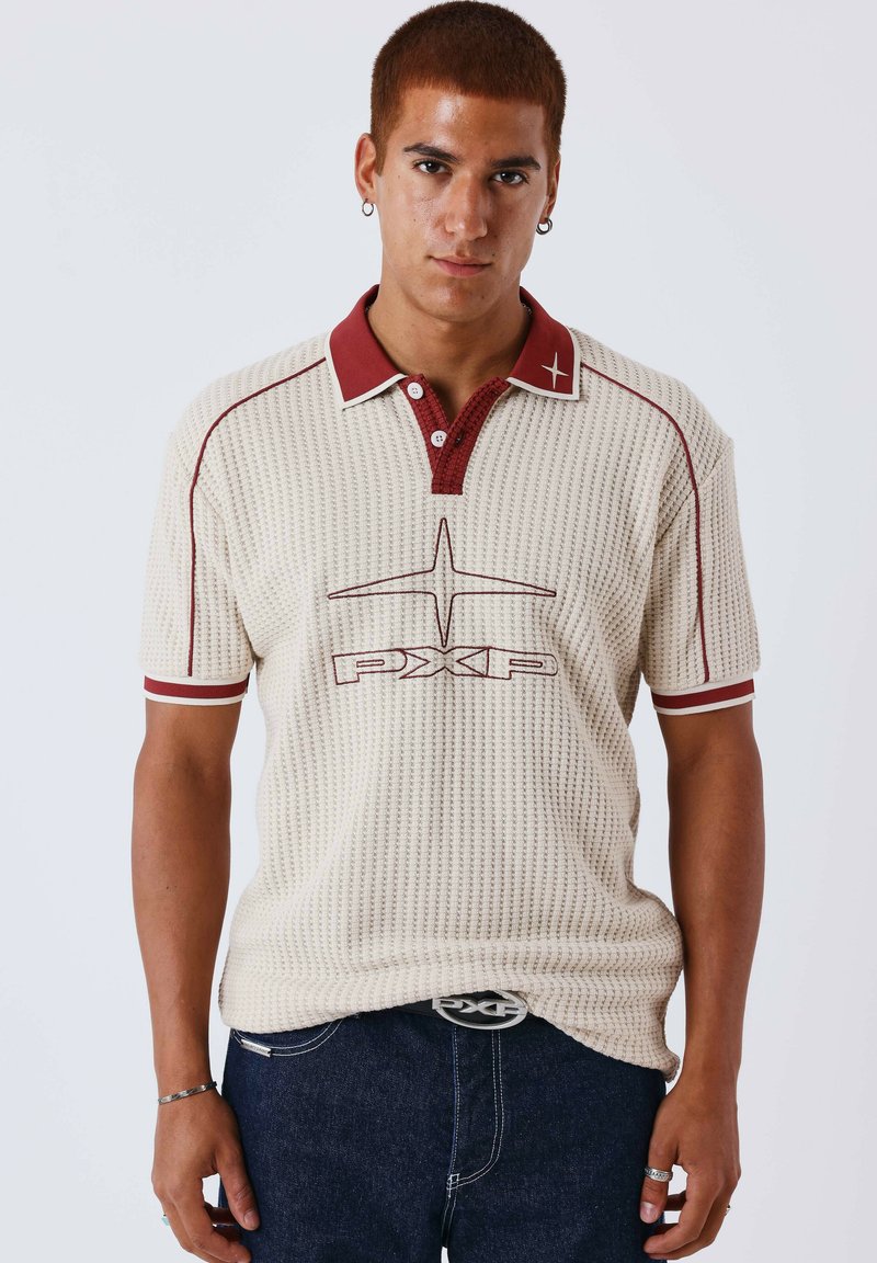 Jeune homme aux cheveux courts portant un polo crème texturé avec col rouge et logo, jean foncé et boucles d'oreilles argentées en forme d'anneau, sur un fond uni.