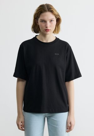 Ung kvinde med kort hår iført en sort BOSS T-shirt og lyseblå jeans, stående mod en ensfarvet lys baggrund.