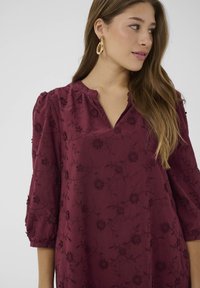 Burgundy blus med 3/4 ärmar, v-ringning, blommig broderi och texturerad väv. Har små blommönster och rynkade ärmdetaljer.