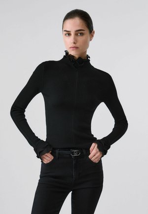 Femme portant un pull noir côtelé ajusté avec un col et des poignets à volants, associé à un jean noir et une ceinture noire, debout devant un fond uni.
