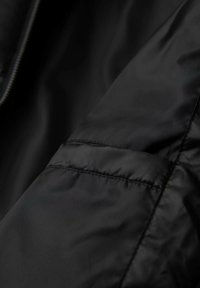 Tessuto della giacca imbottita nera, con una texture liscia caratterizzata da cuciture ben visibili e un dettaglio con cerniera lungo la cucitura. Vista ravvicinata.