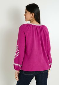 Blouse magenta à manches longues avec col froncé, accents brodés blancs sur les manches et tissu texturé. Associée à un jean foncé.
