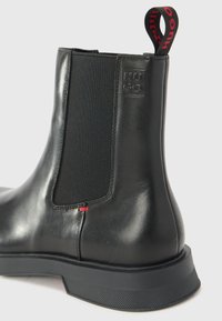 Black leather ankle boot with elastic side panels, textured rubber sole, red accent stitching, and a pull tab featuring "HUGO" branding.
