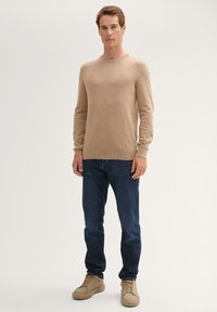 Beige trui van een zachte, lichte stof met een ronde hals en geribbelde manchetten, gecombineerd met donkere spijkerbroek en beige sneakers.