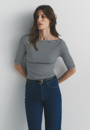 Gestreiftes, figurbetontes Oberteil mit kurzen Ärmeln in Schwarz und Weiß, kombiniert mit hochtaillierter blauer Jeans und einem schwarzen Gürtel mit goldener Schnalle.