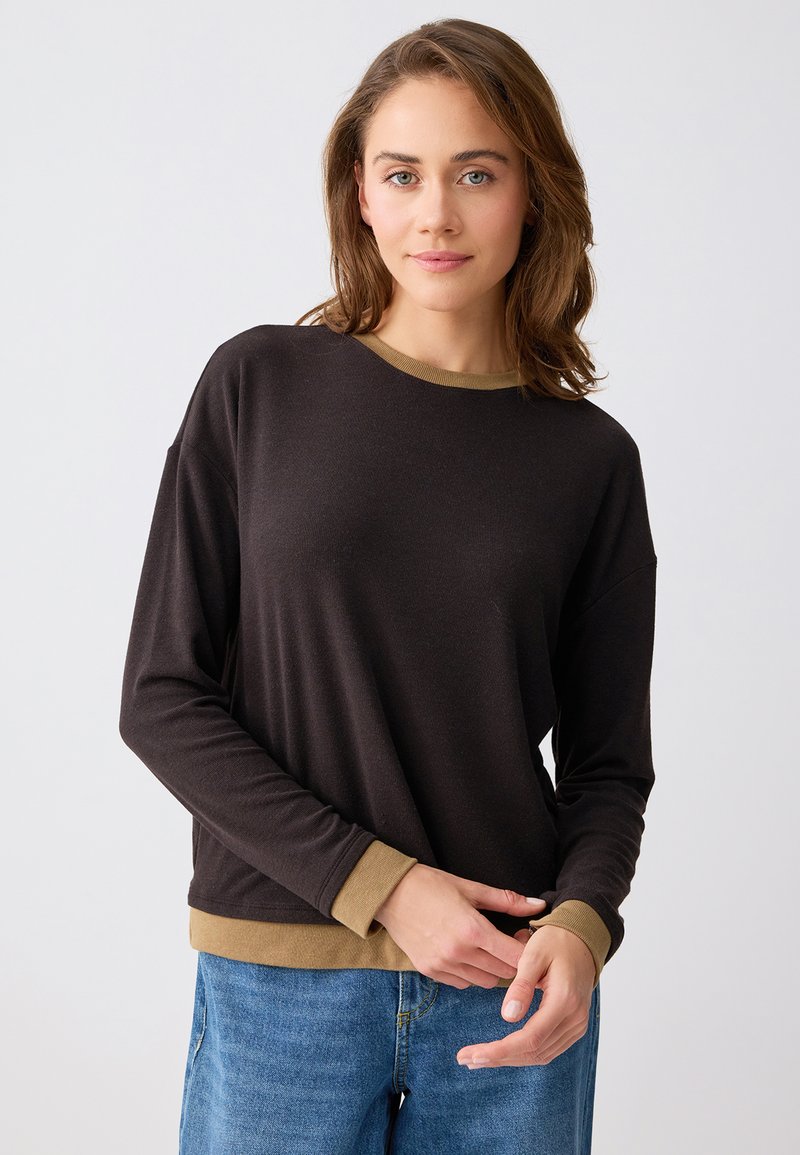 Femme aux cheveux bruns mi-longs portant un haut à manches longues noir avec un bord marron clair et un jean bleu, debout devant un fond blanc.