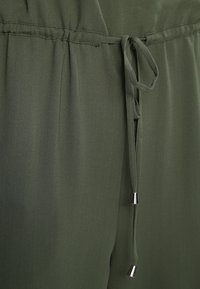 Gros plan sur une combinaison olive, avec une taille froncée dotée d'un cordon de serrage et de pointes métalliques, fabriquée en tissu doux et texturé.