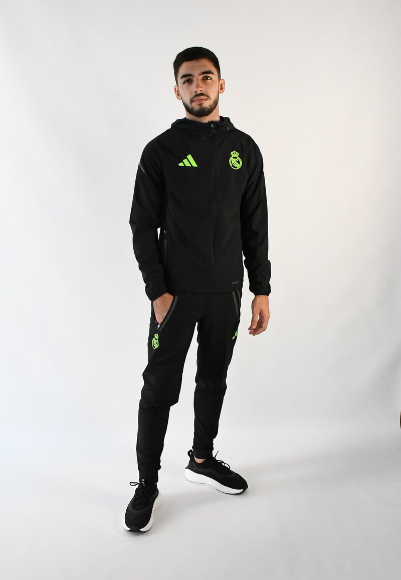 Chándal negro con detalles en verde, que incluye una chaqueta con cremallera y pantalones ajustados. Logo de Adidas y escudo del equipo visibles en ambas prendas.