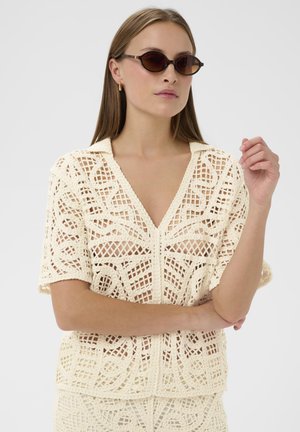 Kreemikrochetsärk sügava V-kaelusega, lühikeste varrukate ja geomeetrilise mustriga. Mudel kannab tumedaid ovaalseid prille.