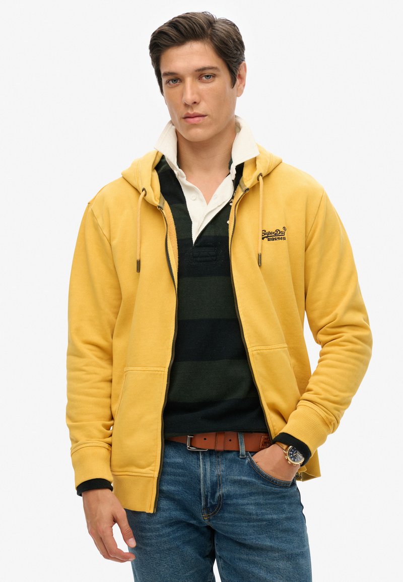 Sudadera con capucha de algodón amarillo con cremallera, que presenta un bolsillo frontal y se lleva sobre una camiseta polo a rayas verde y negra.