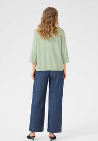 Pull vert clair, texturé, avec des manches trois-quarts, associé à un jean en denim bleu à taille haute et jambes larges, et des talons noirs à dos ouvert.