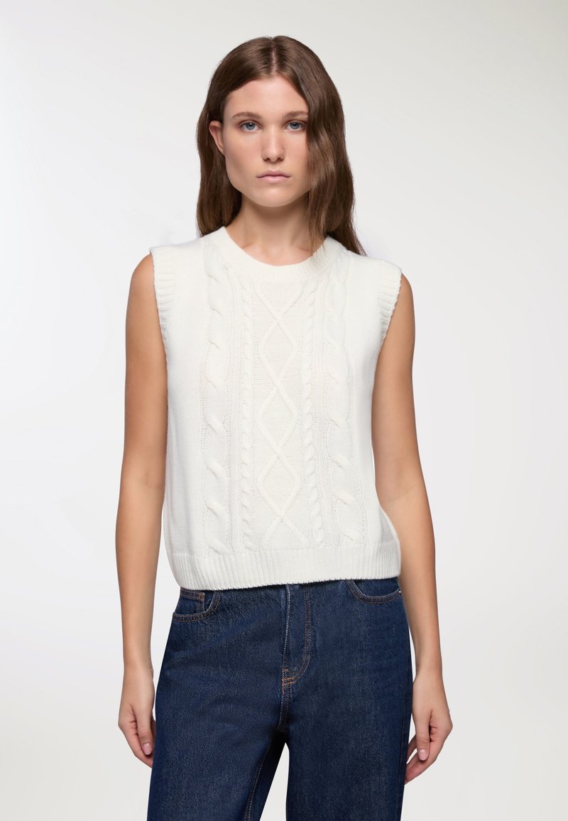 Biała bezrękawnikowa swetrowa bluzka z wzorem w warkocze, okrągłym dekoltem i ściągaczem na dole. W połączeniu z ciemnoniebieskimi dżinsami.