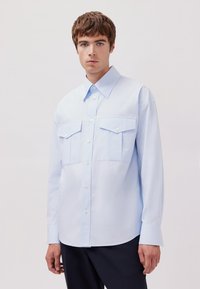 Camicia azzurro chiaro a maniche lunghe realizzata in tessuto morbido, dotata di due tasche sul petto e colletto classico, con chiusura a bottoni sul davanti.