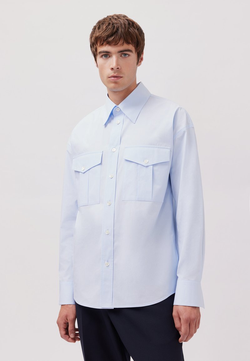 Camicia azzurro chiaro a maniche lunghe realizzata in tessuto morbido, dotata di due tasche sul petto e colletto classico, con chiusura a bottoni sul davanti.