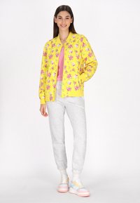 Giacca bomber gialla con motivo floreale rosa, abbinata a una maglietta rosa e pantaloni della tuta grigi. Le calzature presentano colori pastello con un design chunky.