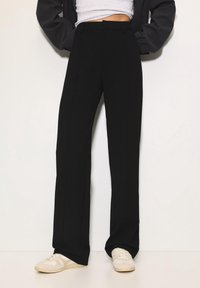 Personne portant un pantalon noir taille haute à jambes droites, un haut court blanc, une veste noire et des baskets blanches, debout devant un fond blanc.