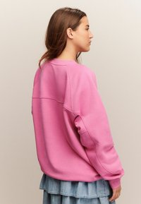 Kobieta z długimi brązowymi włosami, ubrana w oversize'owy różowy sweter i warstwową spódnicę dżinsową, zwrócona w prawo na jednolitym tle.
