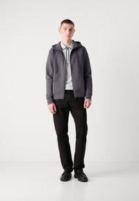Sudadera gris con cremallera de textura suave, combinada con una camiseta polo blanca con rayas negras, pantalones negros ajustados y zapatos negros.
