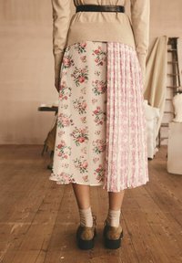 Blumen-Midirock mit einer cremefarbenen Basis, auf einer Seite mit rosa Blumen und auf der anderen Seite mit einem plissierten rosa Abschnitt. Braune Absatzschuhe und Socken.