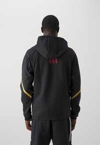 Svart huvtröja med gula och röda detaljer; har vadderade axlar, en mjuk textur och en rak design.