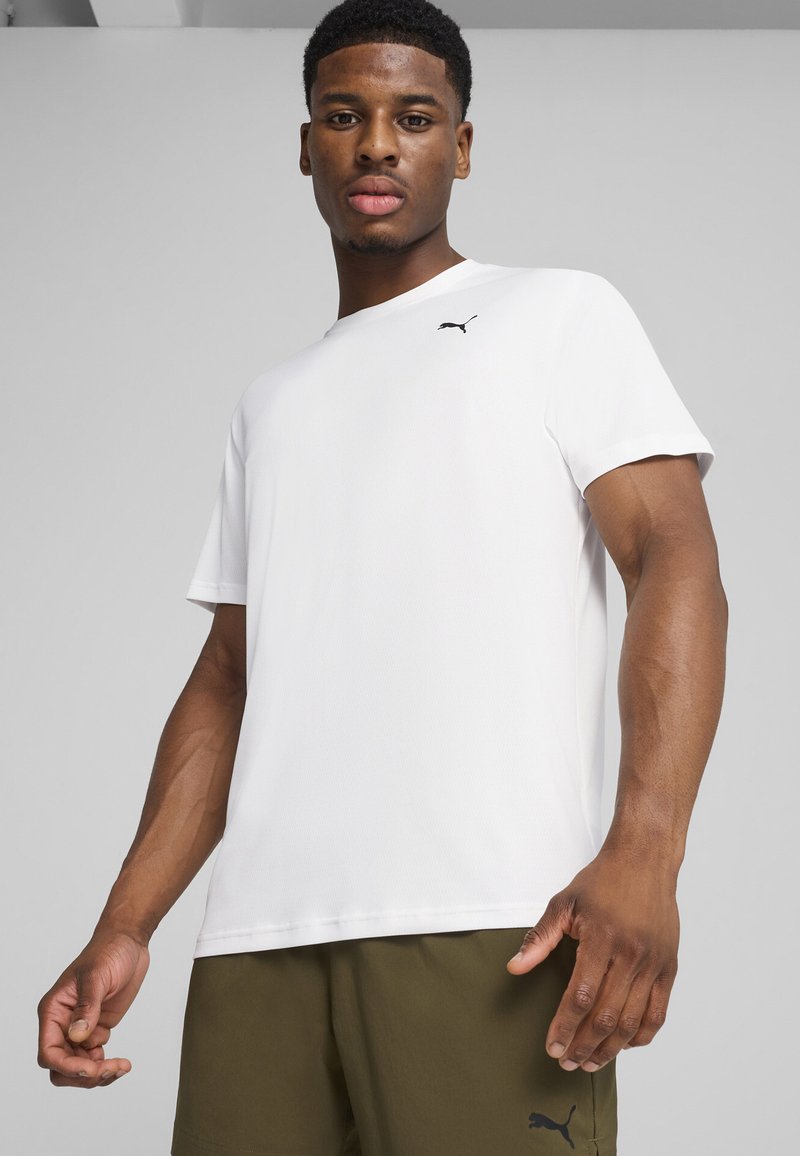 Jeune homme portant un T-shirt blanc Puma et un short Puma vert olive, debout devant un fond gris clair uni.