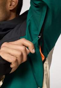 Casaco verde e preto com uma superfície texturizada; apresenta um forro laranja e um zíper visível. Mão segurando o puxador do zíper.