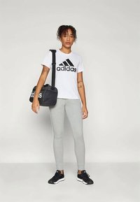 Balti adidas marškiniai su juodu logotipu, pilkos prigludusios kelnės ir juodi sportiniai bateliai. Laiko juodą sportinį krepšį su baltu adidas prekiniu ženklu.