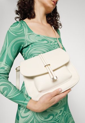 Sac en cuir texturé couleur crème avec fermeture à rabat magnétique et quincaillerie dorée, tenu contre une robe verte à motifs.