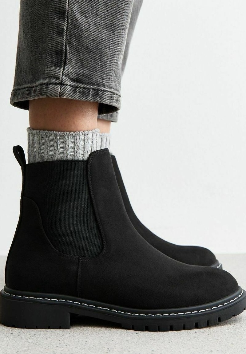 Personne portant des bottines Chelsea en suède noir à semelles épaisses, des chaussettes en laine grises tricotées et un pantalon en denim gris retroussé, debout sur un sol clair.