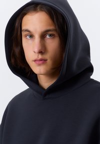 Sweatshirt à capuche noire en matériau doux, avec une coupe décontractée. La capuche est doublée et il n'y a pas de logos ni de motifs visibles.