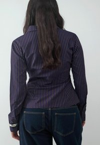 Hemd mit Button-down-Kragen in einem senfgelben und orangefarbenen vertikalen Streifenmuster, langen Ärmeln und taillierter Passform. Getragen mit dunkelblauen Jeans.