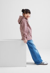 Light pink hooded jacket with a front zipper, paired with blue wide-leg jeans and gray sneakers with black stripes, sitting on a white block.