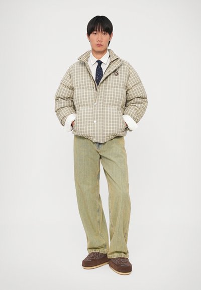 Jakna s pufovima u bež i smeškastom karo uzorku, sa visokim ovratnikom i belim manžetama. Model nosi zelene prugaste pantalone i smeđe cipele.