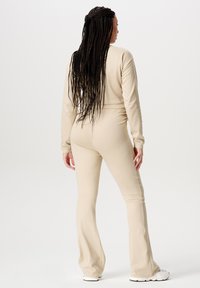 Beige jumpsuit met lange mouwen, een aansluitende bovenkant en uitlopende broekspijpen, gemaakt van zachte stof. Heeft lange gevlochten haren en witte sneakers.