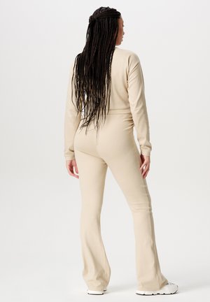 Beige Langarm-Jumpsuit mit tailliertem Oberteil und ausgestellten Beinen, aus weichem Stoff gefertigt. Merkmale sind lange geflochtene Haare und weiße Sneaker.