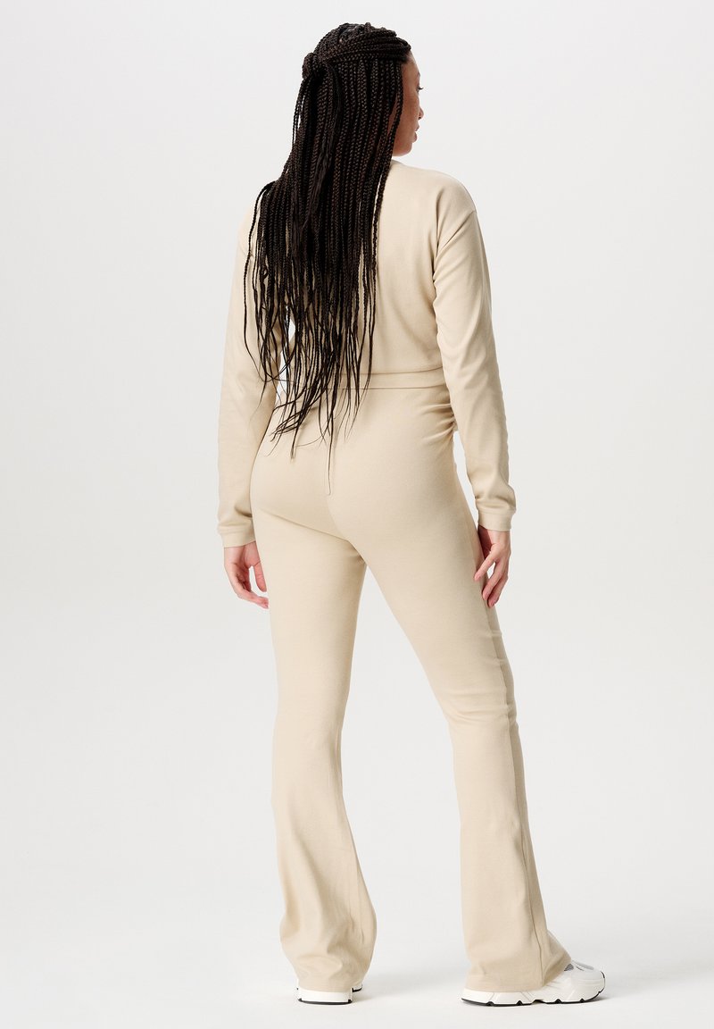 Beige jumpsuit met lange mouwen, een aansluitende bovenkant en uitlopende broekspijpen, gemaakt van zachte stof. Heeft lange gevlochten haren en witte sneakers.