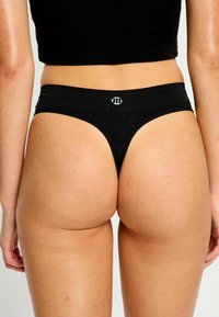 Femme portant un string noir et un crop top noir, vue de dos contre un fond clair.