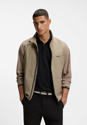 Mężczyzna ubrany w beżową kurtkę zapinaną na zamek, pod którą ma czarną koszulę z kołnierzykiem, czarne spodnie oraz dwukolorowy pasek, stoi z rękami w kieszeniach na białym tle.