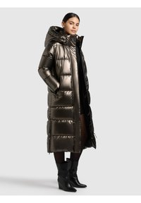 Žena u dugom metalik pufastom kaputu s kapuljačom, crnoj haljini, hulahopkama i crnim gležnjačama na klinastoj peti, stoji ispred jednobojne pozadine.